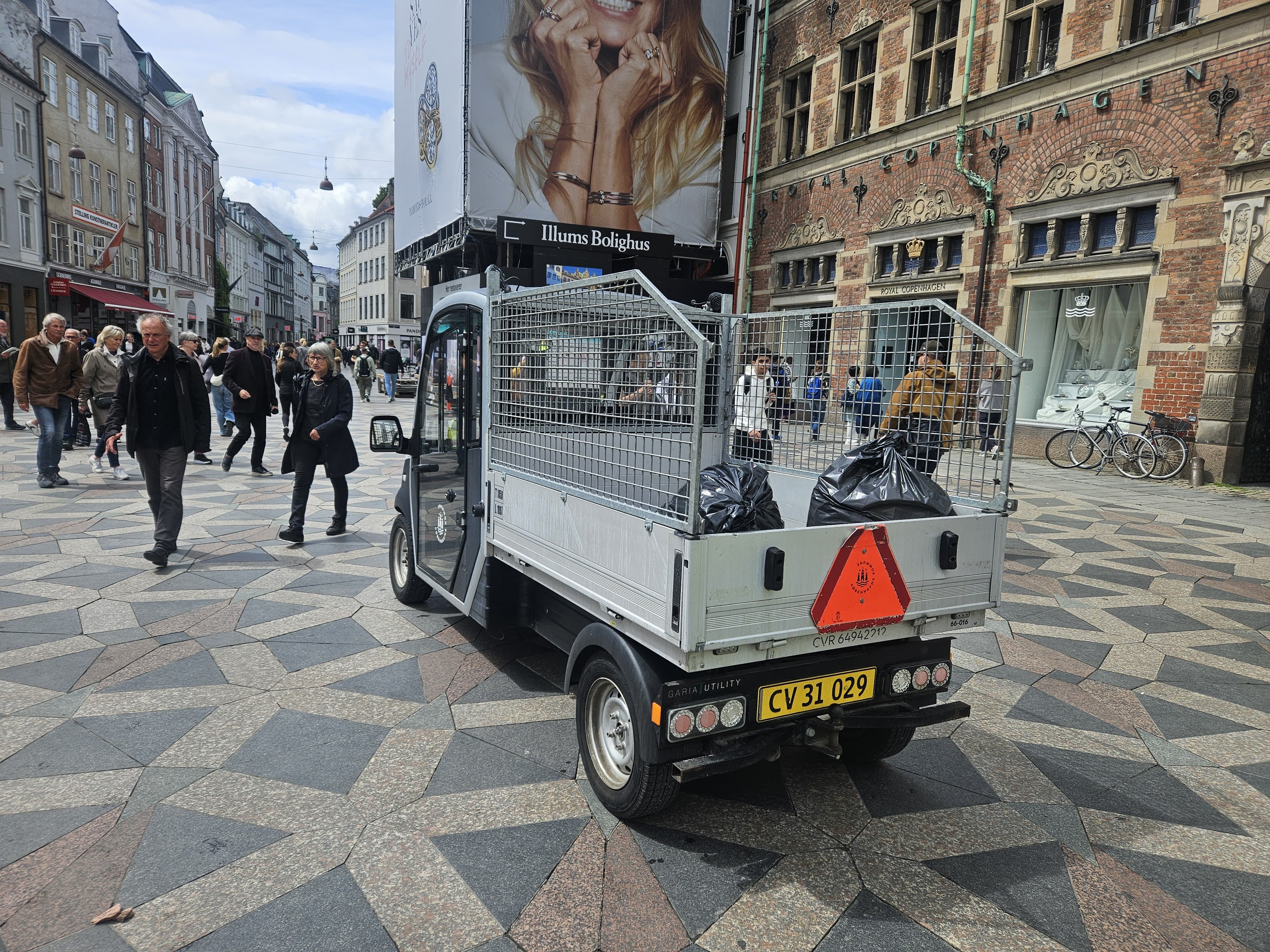 Foto de um pequeno caminhão de lixo branco estacionado em uma rua de paralelepípedos com padrão geométrico. O caminhão tem uma carroceria de grade metálica na traseira, onde duas sacolas de lixo pretas são visíveis. Na parte traseira do caminhão, há um triângulo de sinalização vermelho e uma placa com a inscrição "CV 31 029". Ao fundo, pessoas caminham pela rua e edifícios de tijolos com detalhes arquitetônicos clássicos são visíveis. A foto foi tirada em um ângulo baixo, mostrando a parte traseira do caminhão em destaque.